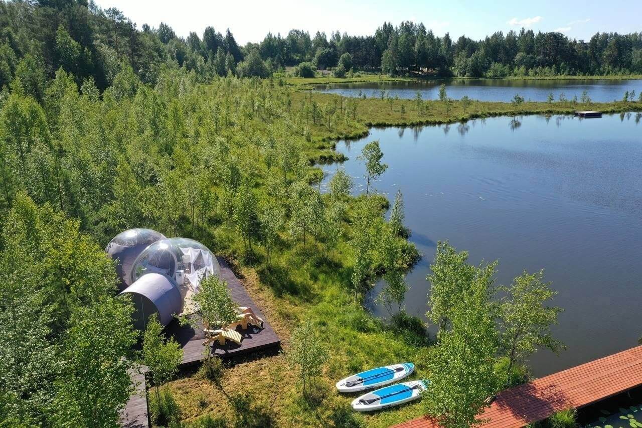 Панорамный вид: густой зеленый лес, берег озера, купальные домики, САП-борды и деревянный пирс.